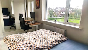 A bedroom with a bed, a desk with a computer, a chair, and a window with a view of houses. - Room-spacious Double Room With Garden & (Rathnew)