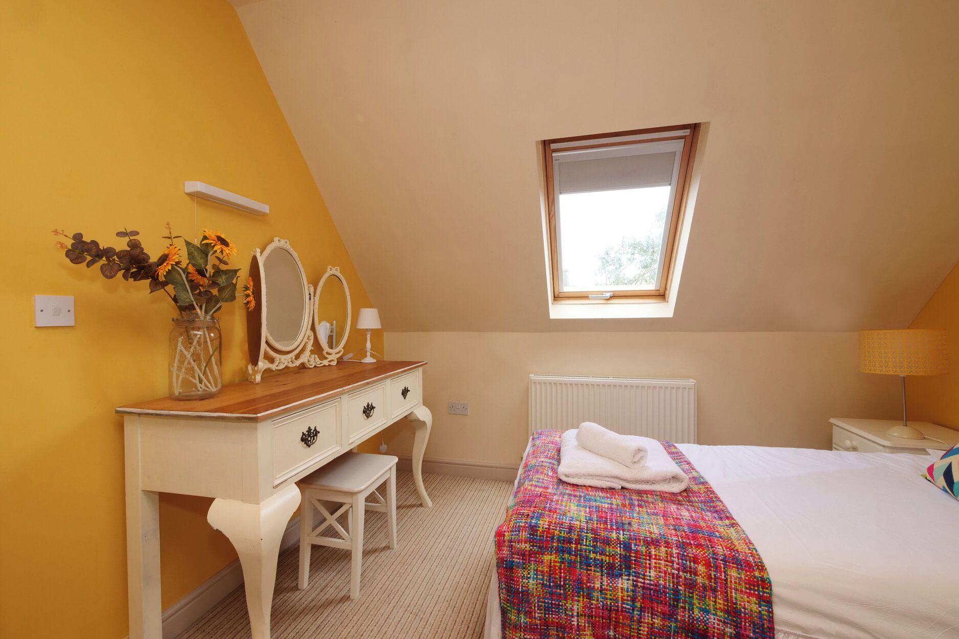 A bedroom with a sloped ceiling, a window, a radiator, a bed with a colorful blanket, a vanity with a mirror and vase, and a small stool.
