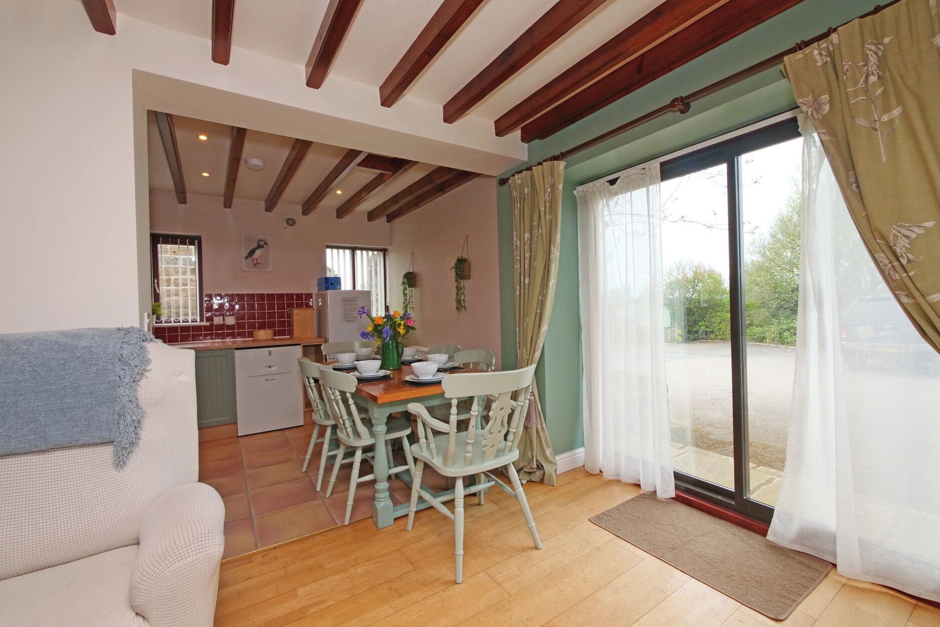 Una cocina acogedora con vigas de madera, un comedor con sillas blancas y un amplio ventanal corredera al exterior.