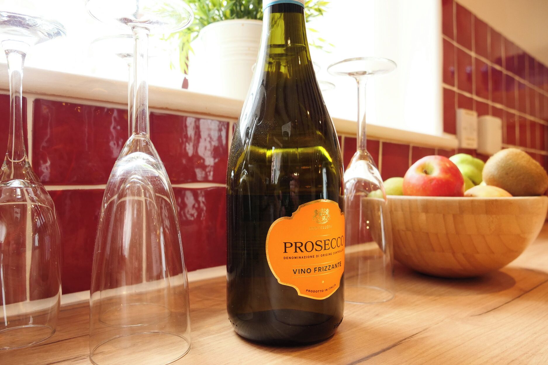 Uma garrafa de vinho Prosecco com etiqueta, rodeada por copos de vinho vazios e uma taça de fruta sobre uma superfície de madeira.