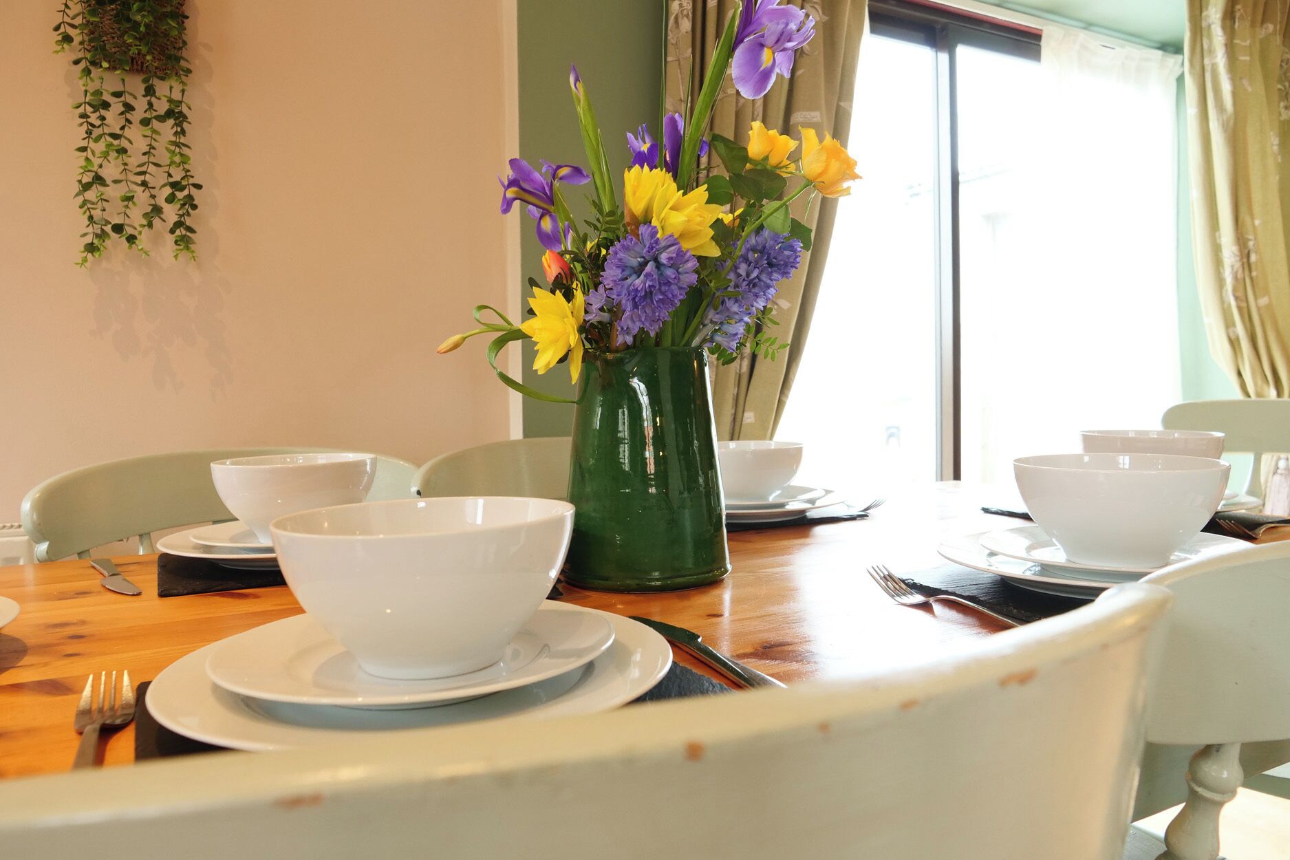 A dining table with white bowls and plates, a green vase with flowers, and a window with curtains.