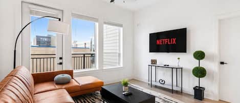 Un salon moderne avec un canapé en cuir, une table basse noire, un téléviseur fixé au mur et un ventilateur de plafond.