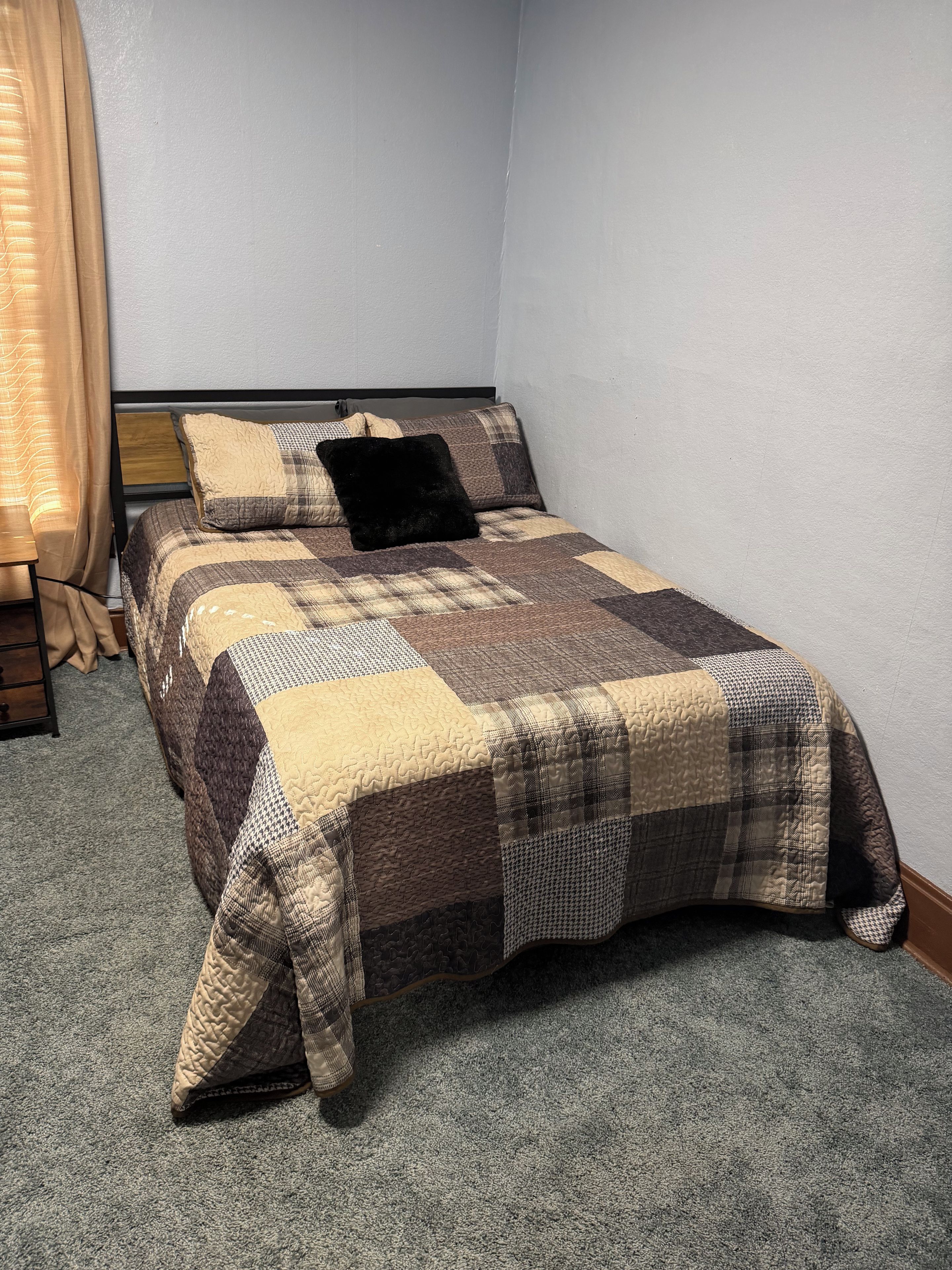 A single bed with a quilted bedspread, a wooden headboard, and a window with curtains.