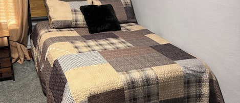 A single bed with a quilted bedspread, a wooden headboard, and a window with curtains.