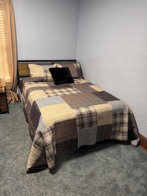 A single bed with a quilted bedspread, a wooden headboard, and a window with curtains.