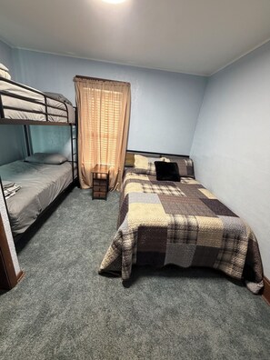 A dormitory room with bunk beds, a single bed, a small wooden chair, and a window with curtains.