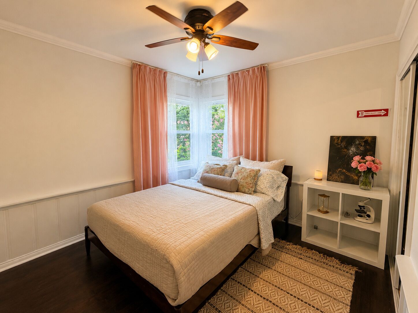 A bedroom with a bed, a ceiling fan, a window with curtains, a bedside table with a lamp, and a shelf with decorative items.