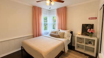 A bedroom with a bed, a ceiling fan, a window with curtains, a bedside table with a lamp, and a shelf with decorative items.