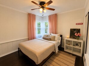 Un dormitorio con cama, ventilador de techo, ventana con cortinas, mesita de noche con lámpara y estante con objetos decorativos.