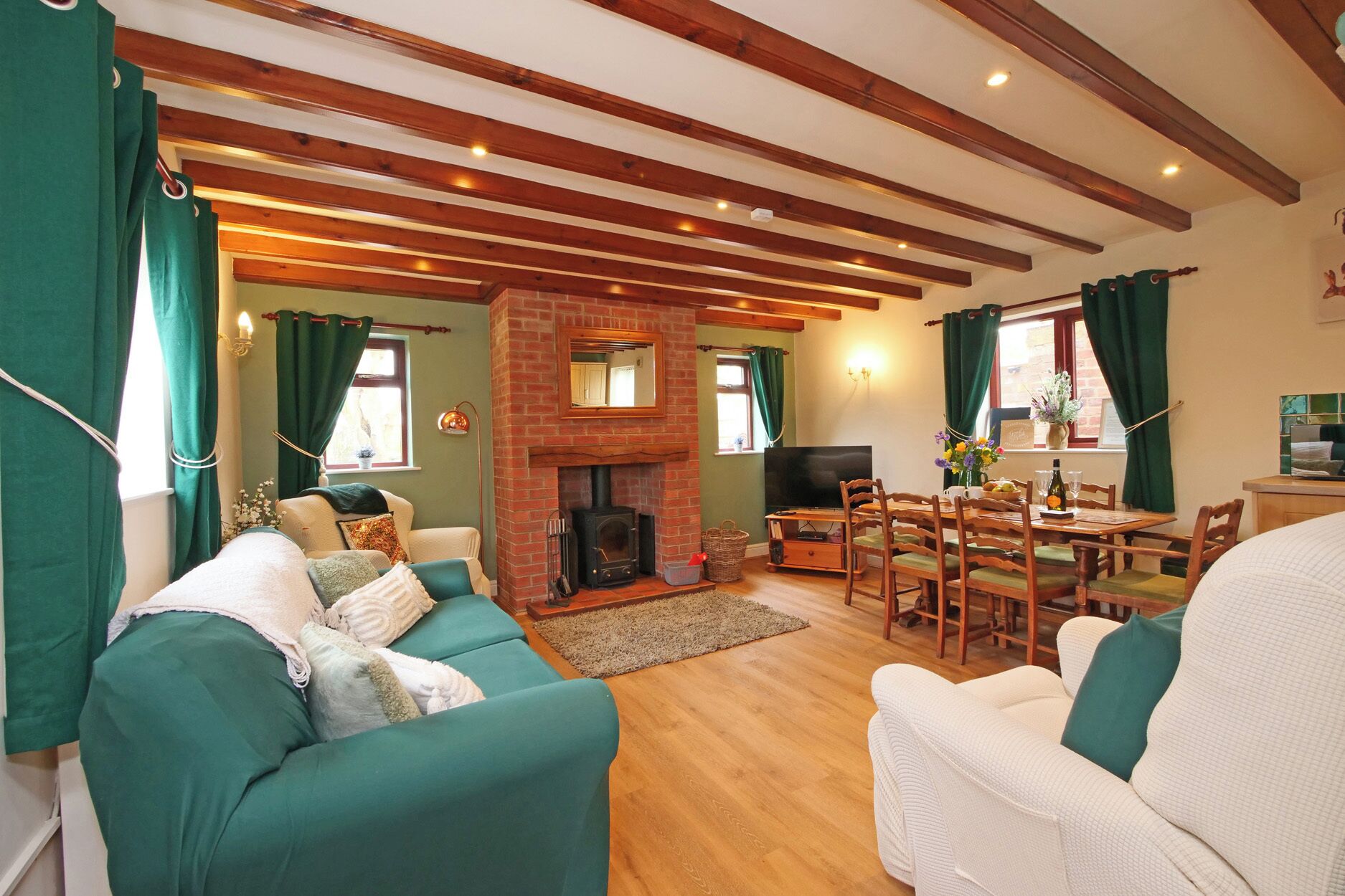 A cozy living room with a fireplace, green curtains, wooden beams, and a dining area with a table and chairs.