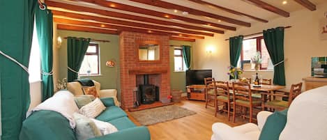 A cozy living room with a fireplace, green curtains, wooden beams, and a dining area with a table and chairs.