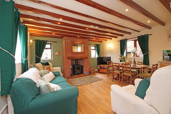 A cozy living room with a fireplace, green curtains, wooden beams, and a dining area with a table and chairs. - Chomley @ Summerfield Farm (Whitby)