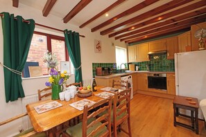 A kitchen with wooden cabinets, a dining table with chairs, a window with curtains, and a brick wall. - Chomley @ Summerfield Farm (Whitby)