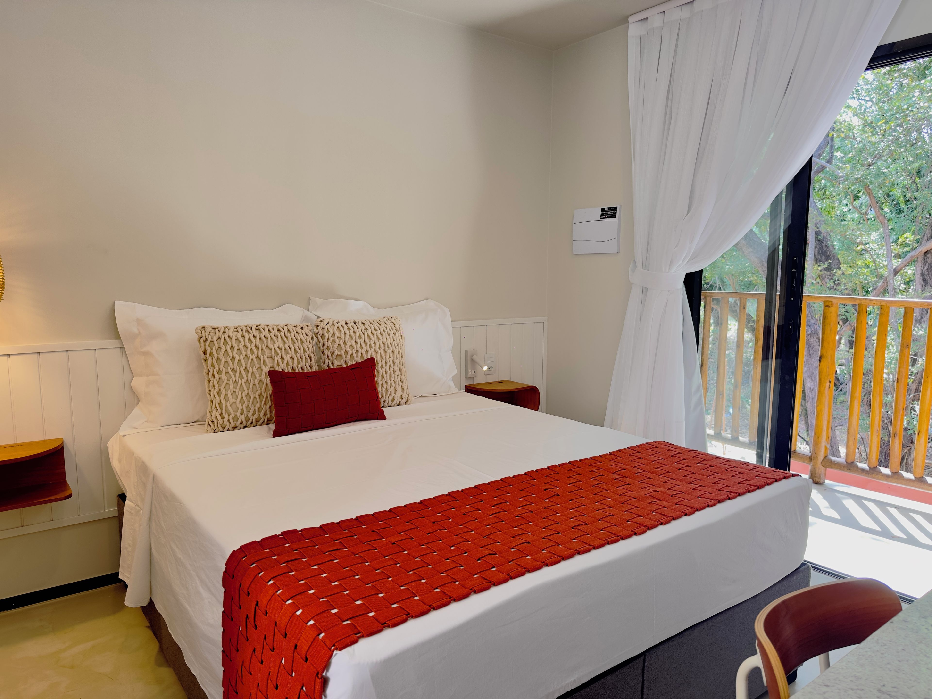 Una habitación de hotel con cama, mesita de noche, una silla roja y vistas al exterior.