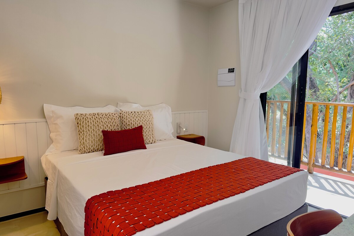 A hotel room with a bed, a nightstand, a red chair, and a view of the outdoors.