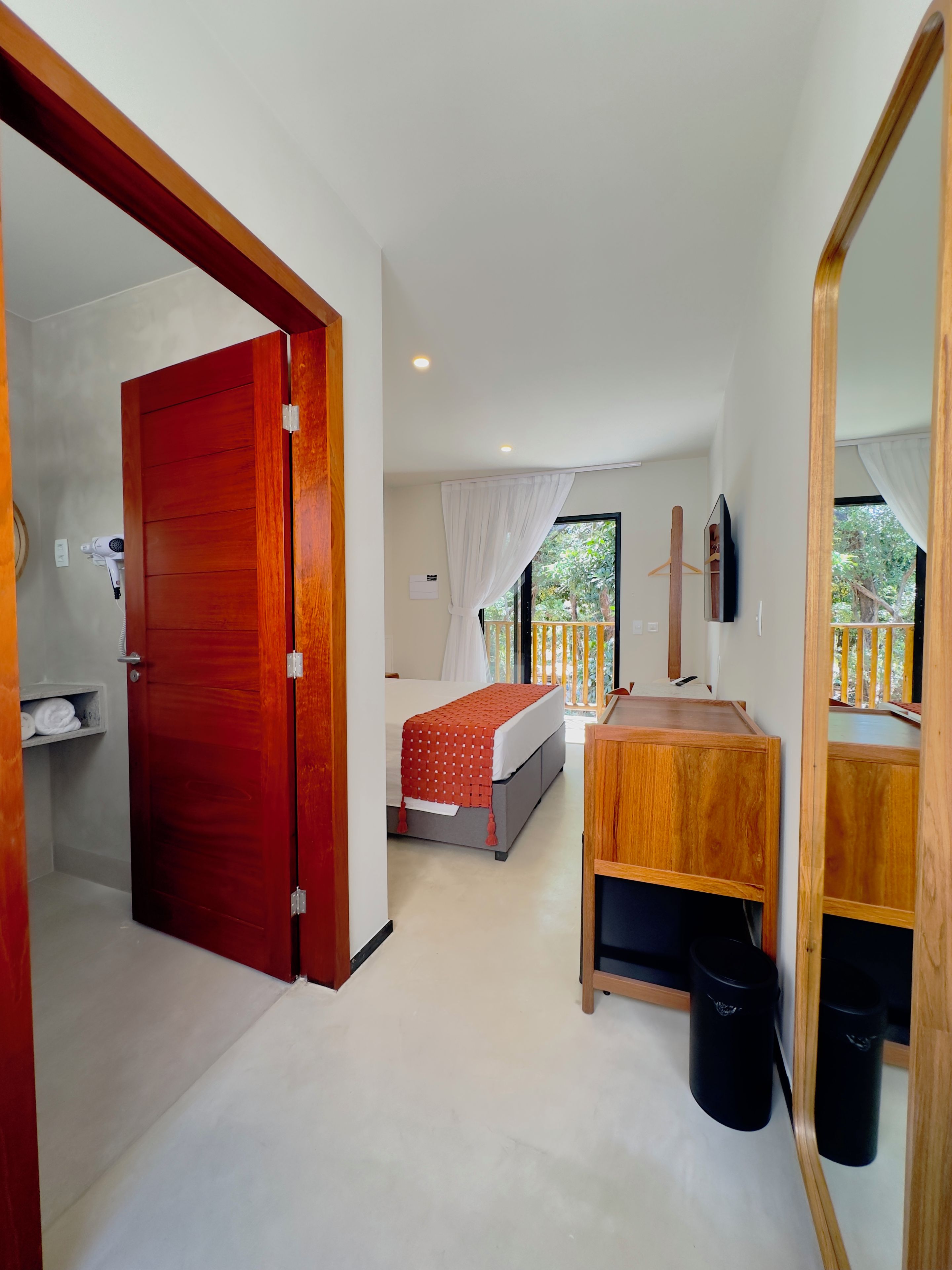 A hotel room with a bed, a wooden desk, a mirror, a TV, and a balcony with a railing.