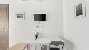 Una habitación con puerta de madera, pared blanca, televisor montado, planta pequeña en maceta, escritorio despejado con una silla y un cubo de basura.