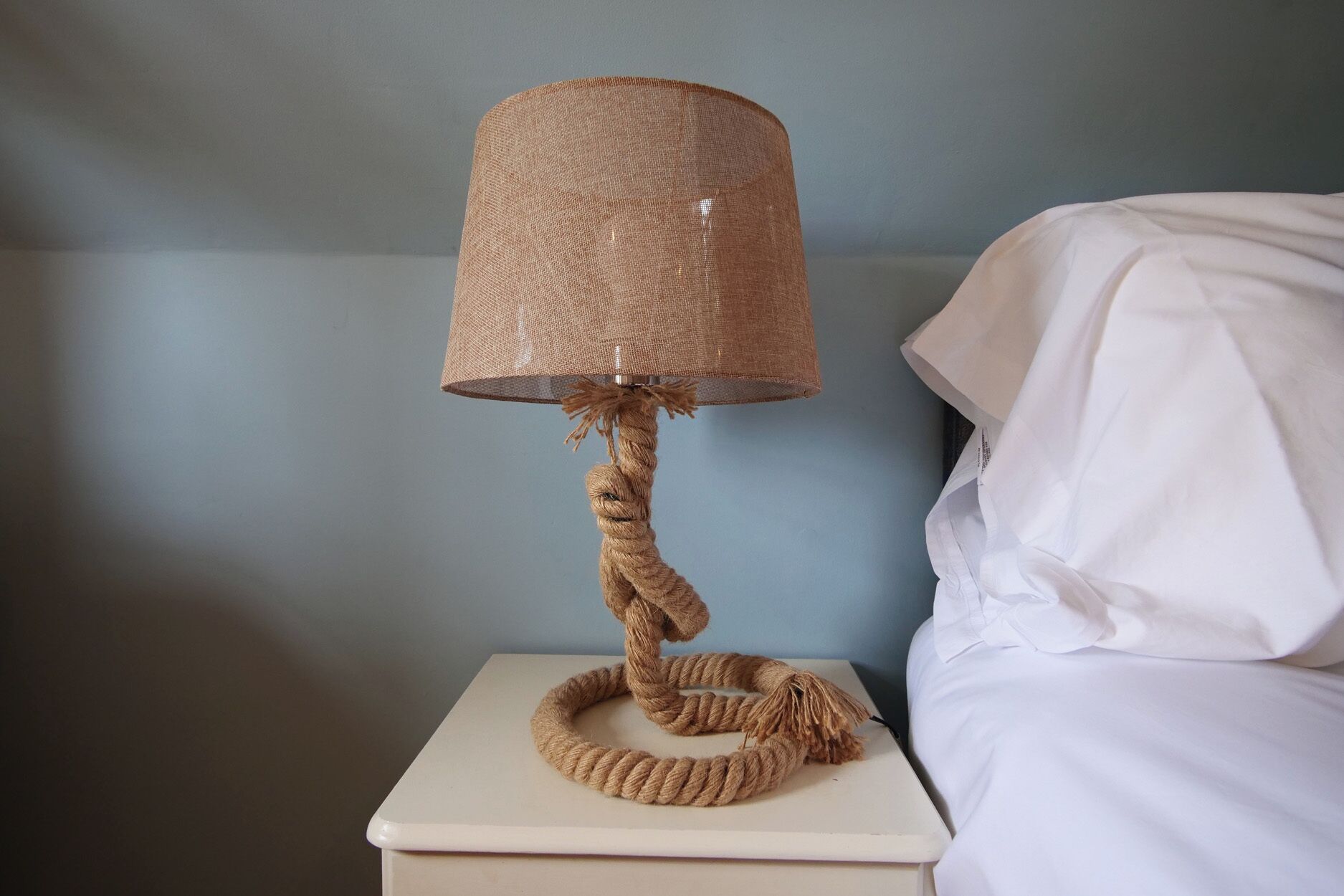 Eine Lampe mit strukturierter Schirm und Kordelbasis auf einem Nachttisch neben einem Bett.
