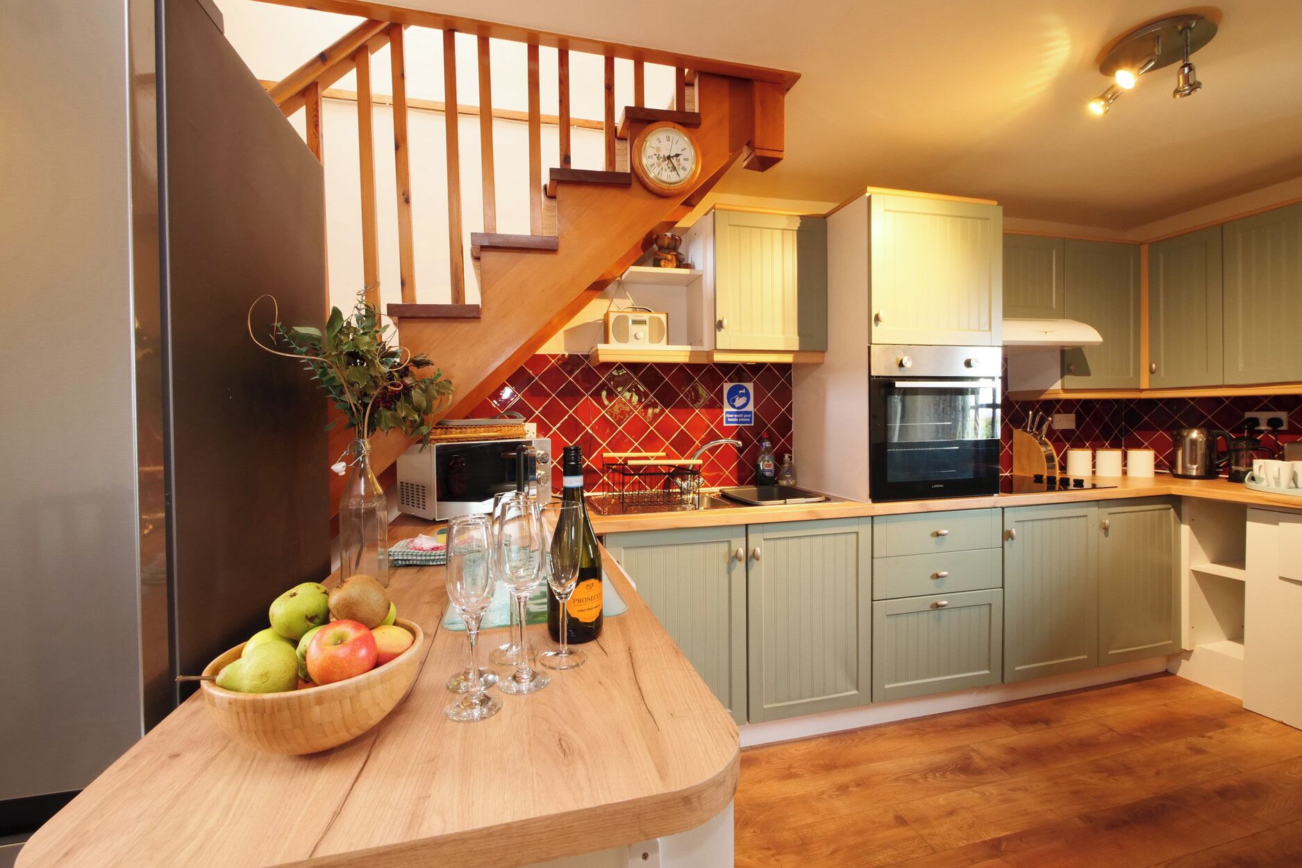 A kitchen with a wooden counter, a bowl of fruit, wine glasses, and a clock on the wall.