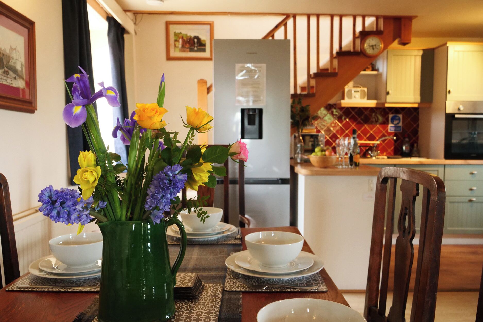 A dining area with a table set for four, a green vase with flowers, a refrigerator, and a kitchen in the background.