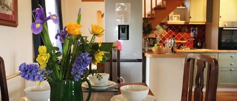 A dining area with a table set for four, a green vase with flowers, a refrigerator, and a kitchen in the background.
