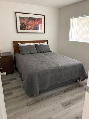 Un dormitorio con cama, una mesita de noche, un cuadro enmarcado en la pared y una ventana con persianas.