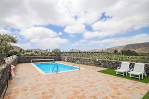 A swimming pool with a stone deck, two white lounge chairs, and a stone wall with plants.