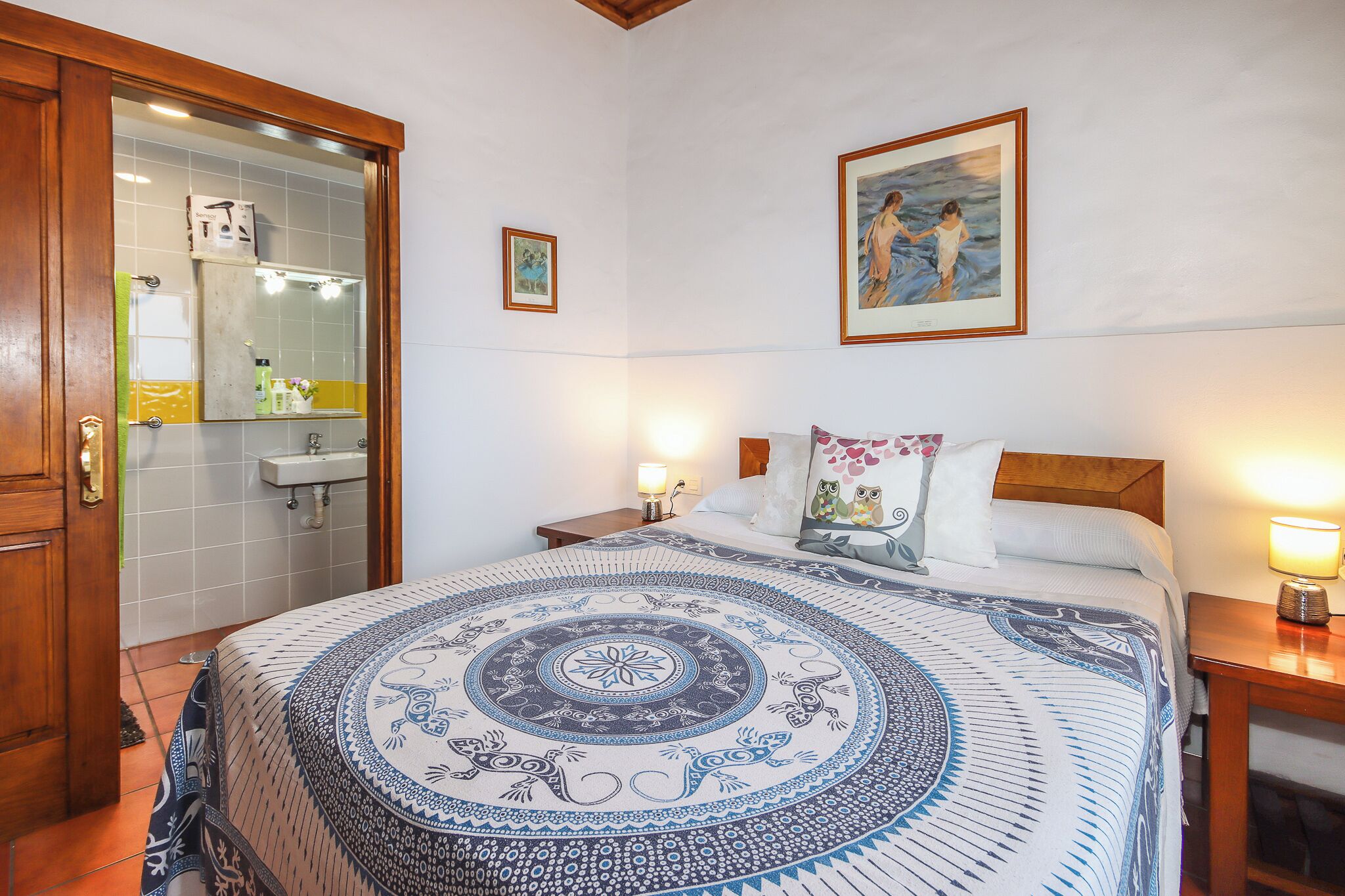 A bedroom with a bed, a nightstand, a bedside lamp, a painting on the wall, and a bathroom with a mirror and sink.
