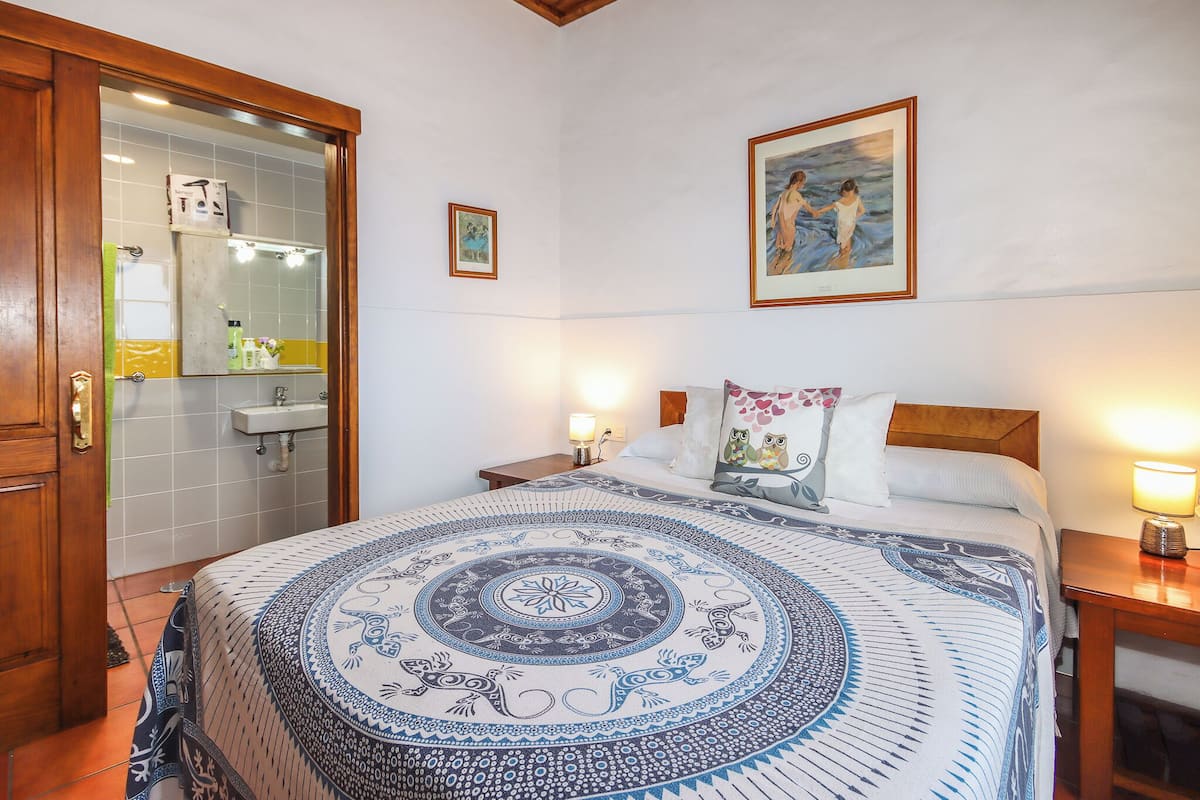 A bedroom with a bed, a nightstand, a bedside lamp, a painting on the wall, and a bathroom with a mirror and sink.
