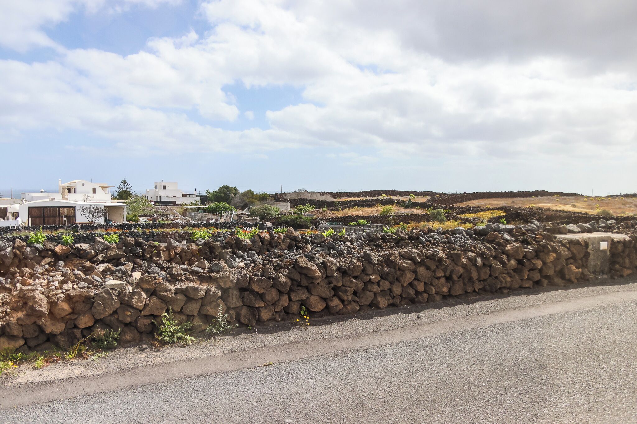 A rural road with a stone wall and buildings in the background.