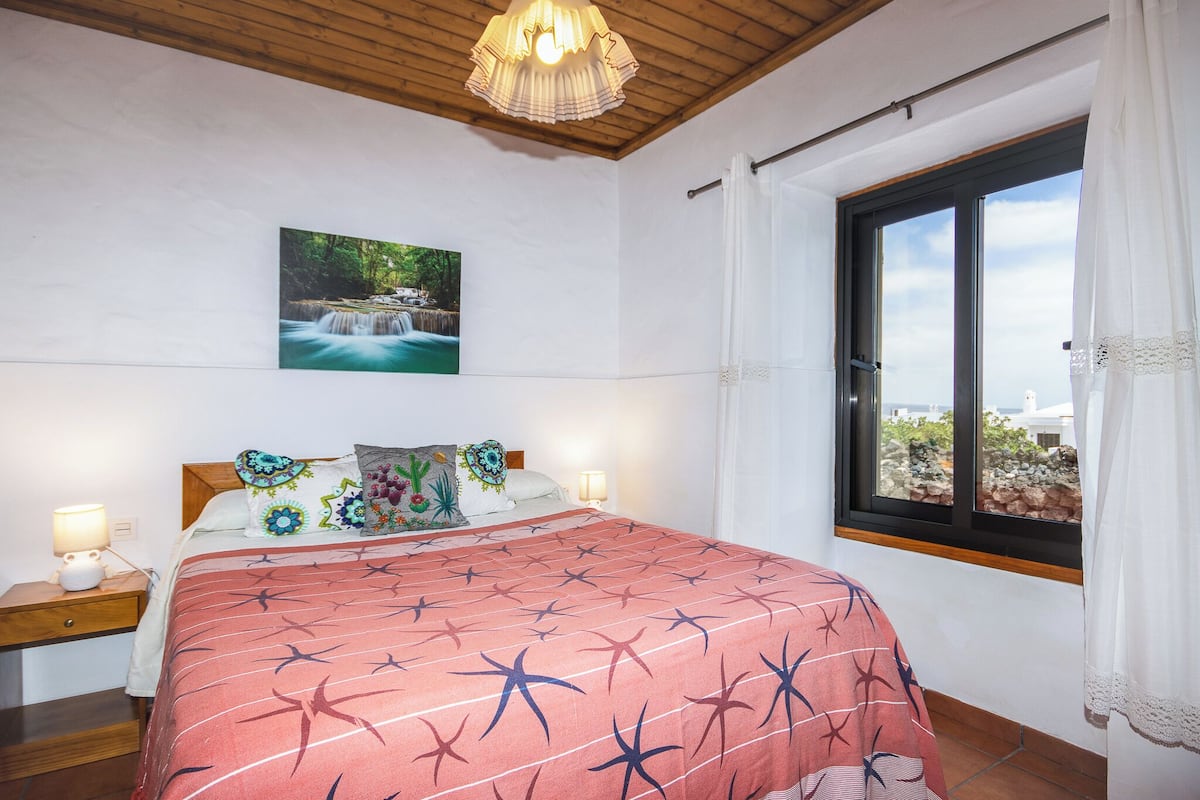 A bedroom with a bed, a nightstand, a window with curtains, a painting on the wall, and a wooden ceiling.
