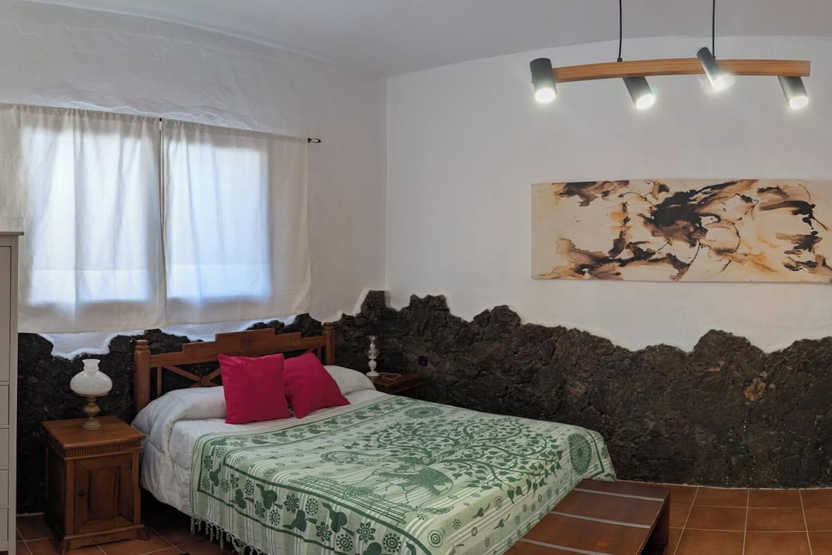 A bedroom with a bed, a nightstand, a dresser, a painting, and a unique stone wall design.