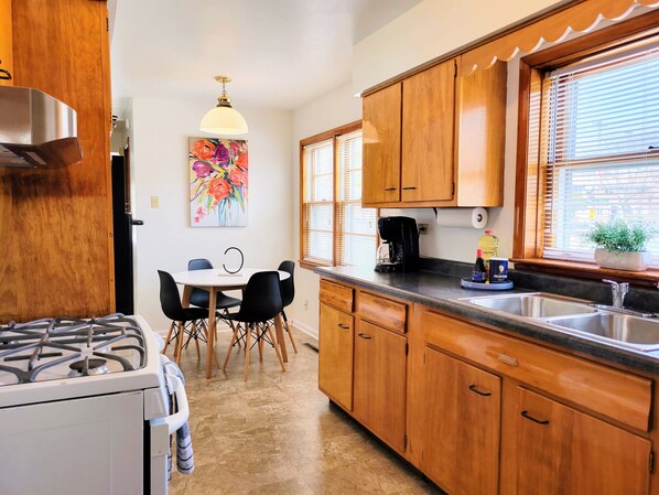 A kitchen with wooden cabinets, a gas stove, a dining table with chairs, a coffee maker, and a window with blinds.