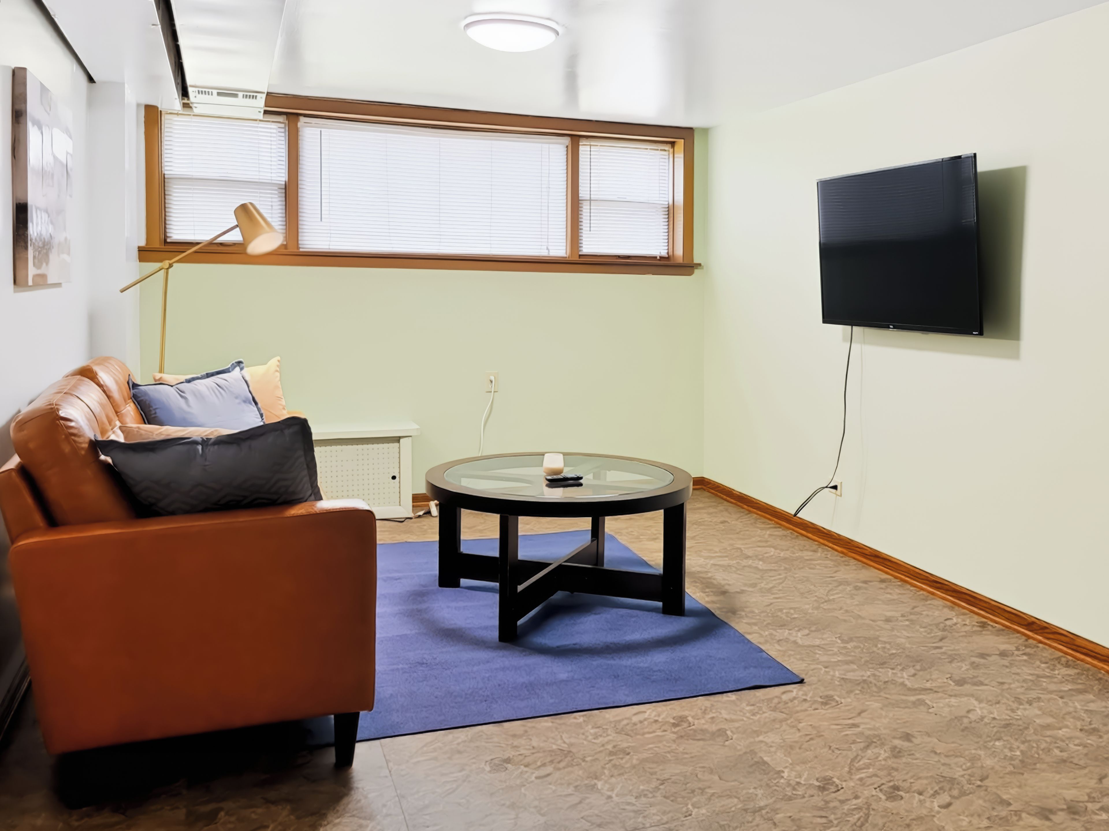 Een gemeubileerde woonkamer met een leren bank, een glazen salontafel, een wandgemonteerde televisie en een raam met jaloezieën.