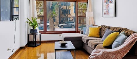 A modern living room with a wooden floor, a brown sofa with cushions, a small table, and a window with a view of the street.