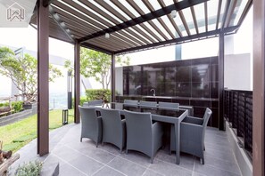 Eine Terrasse im Freien mit einem Tisch aus Glas und Korbstühlen, einer Pergola darüber und Blick auf das Meer.
