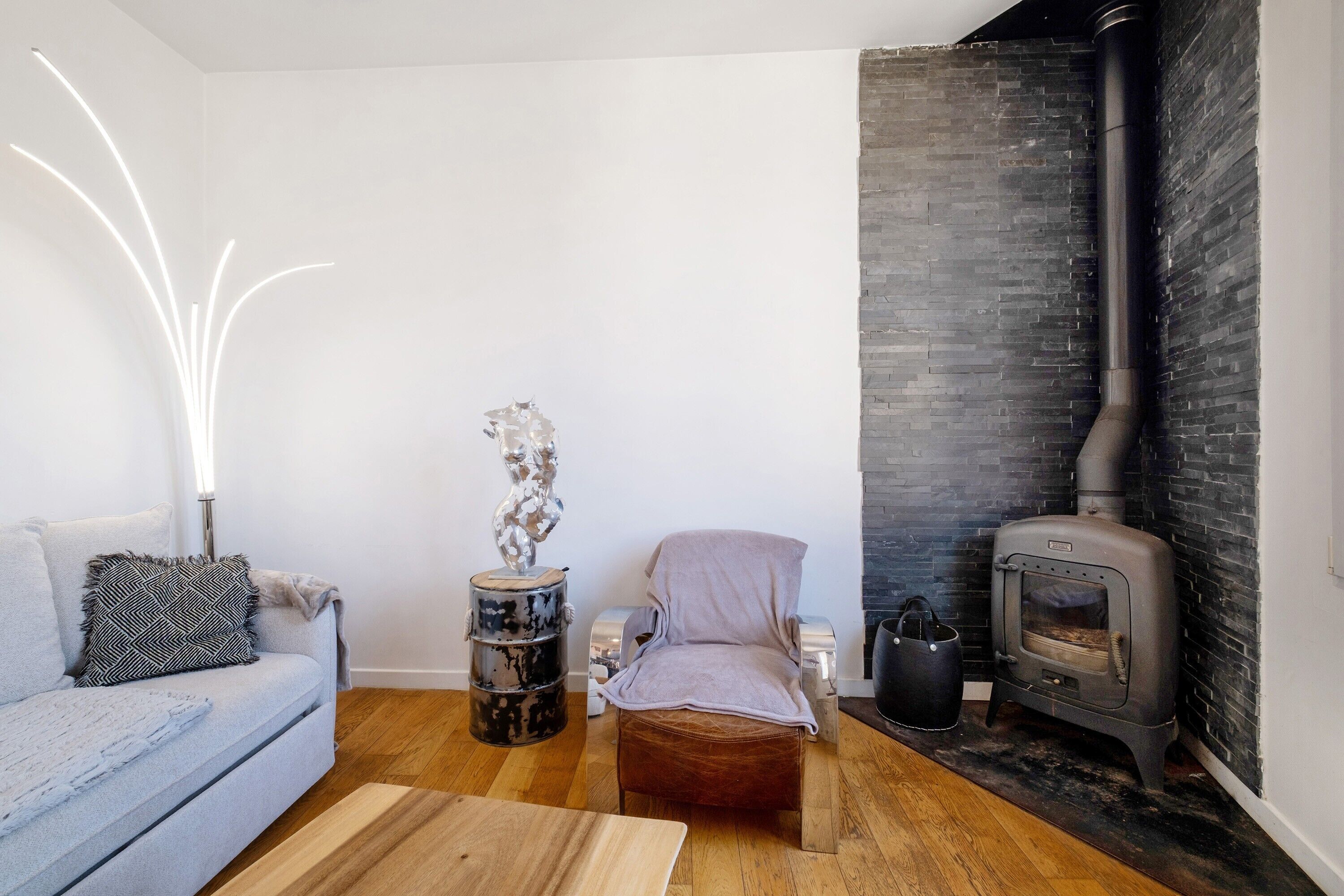 A living room with a sofa, a chair, a wooden coffee table, a decorative sculpture, a black stove, and a white wall with a black brick accent.