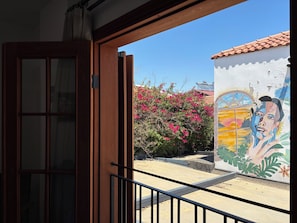 Vista a través de una ventana de madera con baranda metálica, un mural con rostro y paisaje, y flores rosadas al fondo.