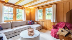 Ein gemütliches Zimmer mit einer Eckcouch, einem runden Hocker, einem roten Karo-Sessel und einem Holztisch mit einer Lampe.