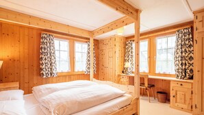 Ein Schlafzimmer einer Holzhütte mit einem Bett, zwei mit Vorhängen versehenen Fenstern, einem Schreibtisch und einem Stuhl.