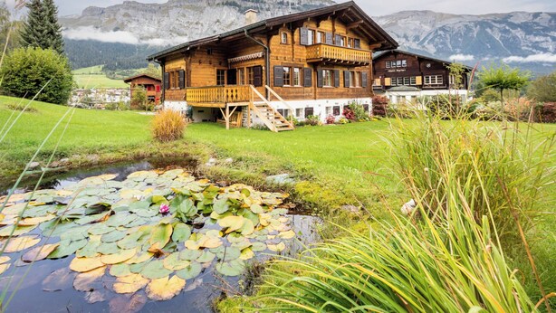 Eine Holzhütte mit Balkon, ein kleiner Teich mit Seerosen und eine Bergkulisse.