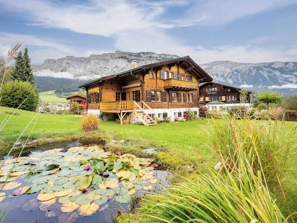 Eine Holzhütte mit Balkon, ein kleiner Teich mit Seerosen und eine Bergkulisse.