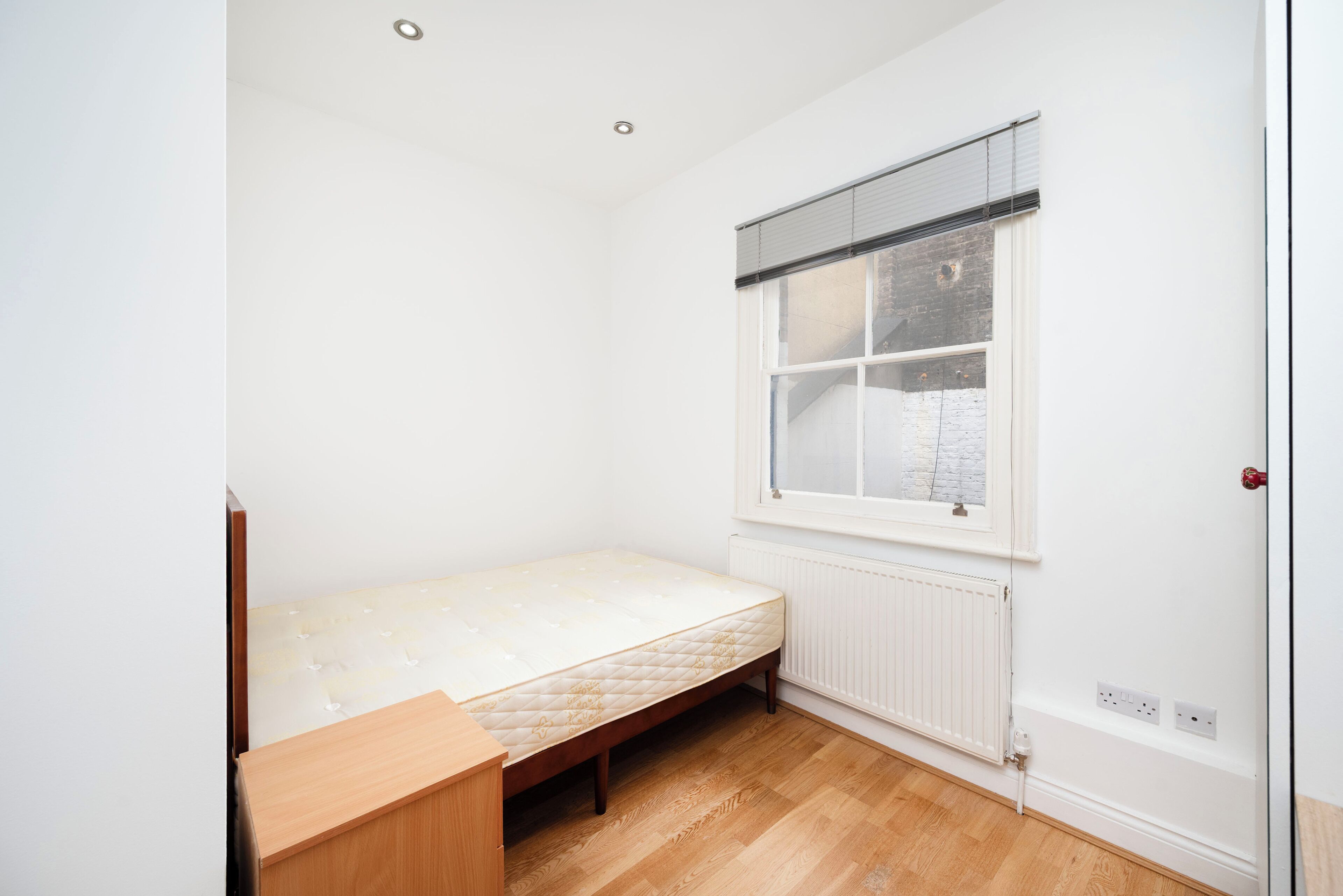 A small, single-bed room with a wooden floor, a window with blinds, a radiator, and a door with a handle.