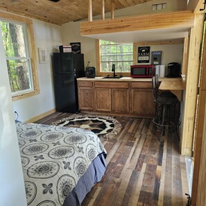 A compact bedroom with a bed, a wooden floor, a kitchen area with a sink, a refrigerator, and a microwave.