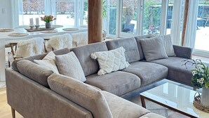 A modern living room with a grey sectional sofa, a glass coffee table, and a dining area with a white table and yellow pendant lights. - 4 Star Holiday Home in Jaegerspris (Jægerspris)