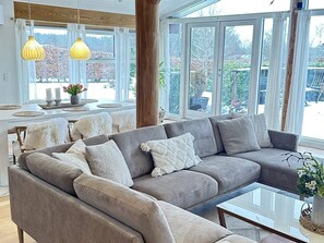 A modern living room with a grey sectional sofa, a glass coffee table, and a dining area with a white table and yellow pendant lights. - 4 Star Holiday Home in Jaegerspris (Jægerspris)