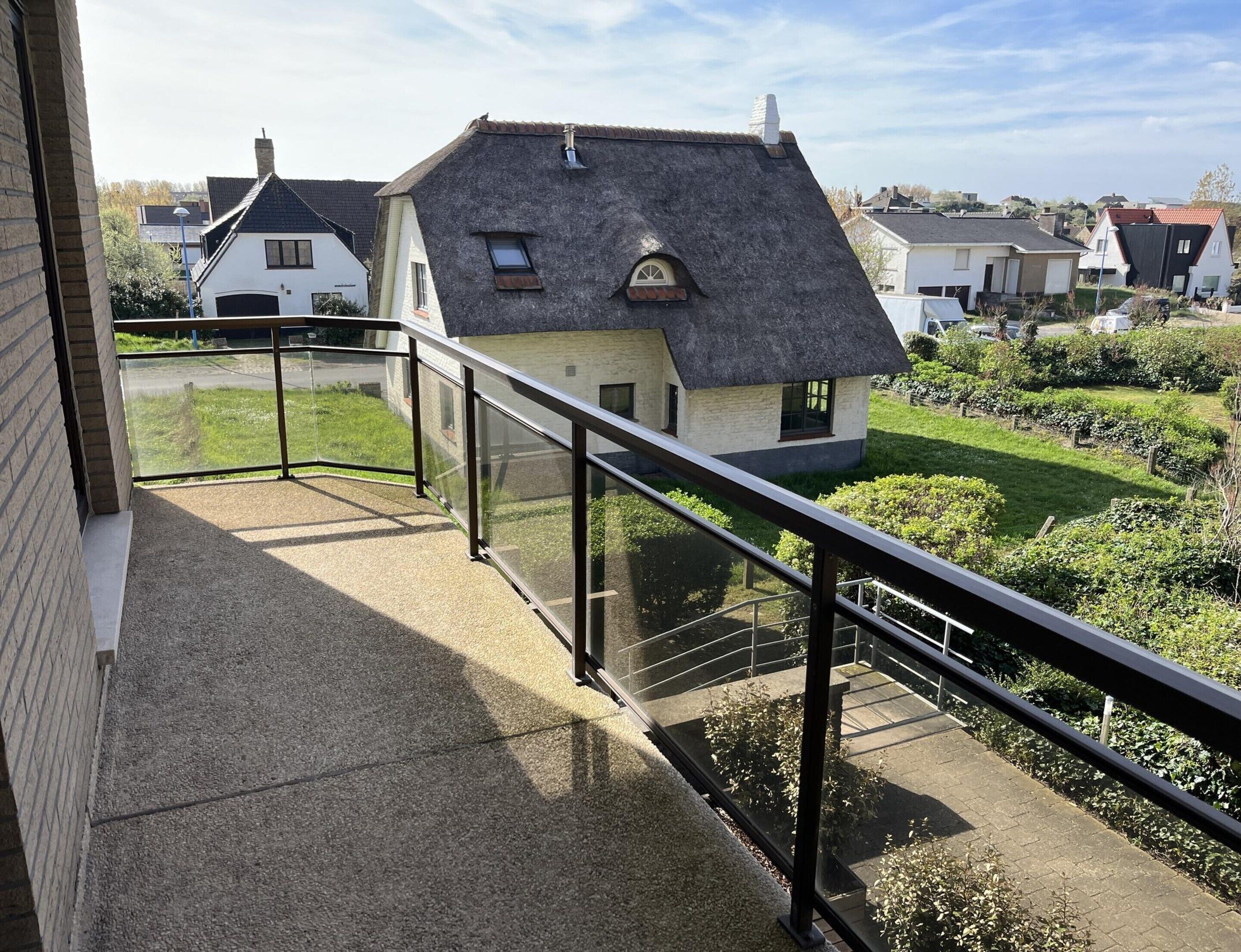 Un balcon doté d’une balustrade en verre, donnant sur un jardin et une maison à toiture de chaume.
