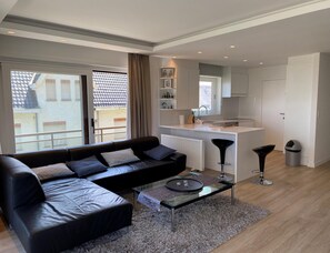 Ein modernes Wohnzimmer mit einer schwarzen Ledersofa, einem Glastisch und Blick auf eine Skyline durch große Fenster.