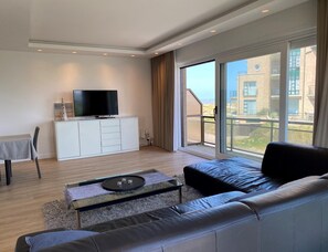Ein modernes Wohnzimmer mit einer Couch, einem Fernseher auf einem Schrank und einem großen Fenster mit Blick auf Gebäude.