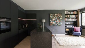 Una cocina moderna con armarios oscuros, una isla de mármol y una zona de comedor con alfombra blanca y una silla azul.